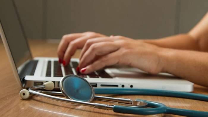 Woman typing on a laptop with a stethoscope next to it