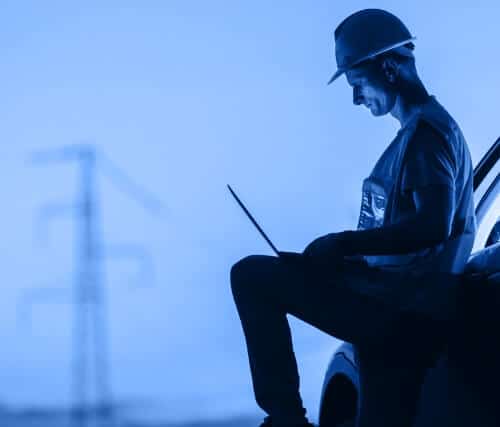 Person wearing a hard hat and vest using a laptop with power lines in the background